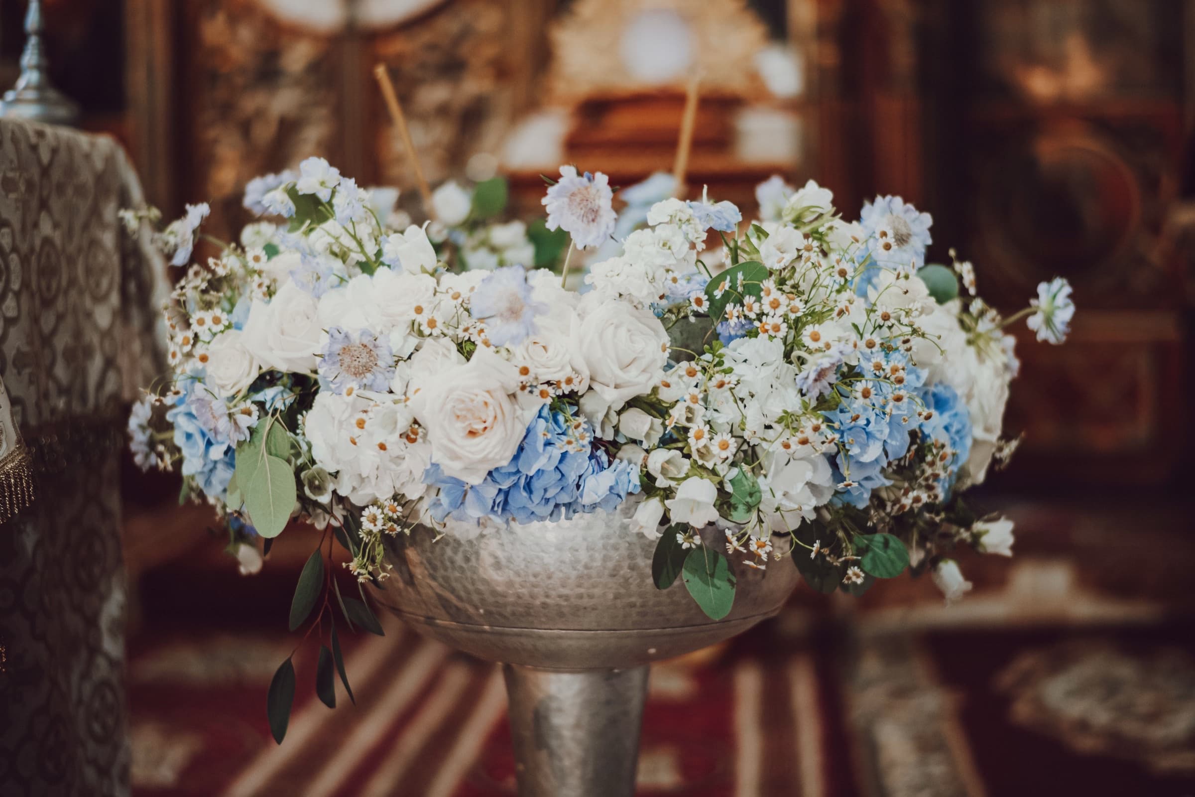 Aranjament floral elegant cu trandafiri albi și hortensii albastre, într-o vază de argint la o ceremonie de botez
